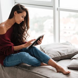 Smiling pretty woman in sweater using tablet computer while sitting on a windowsill at home