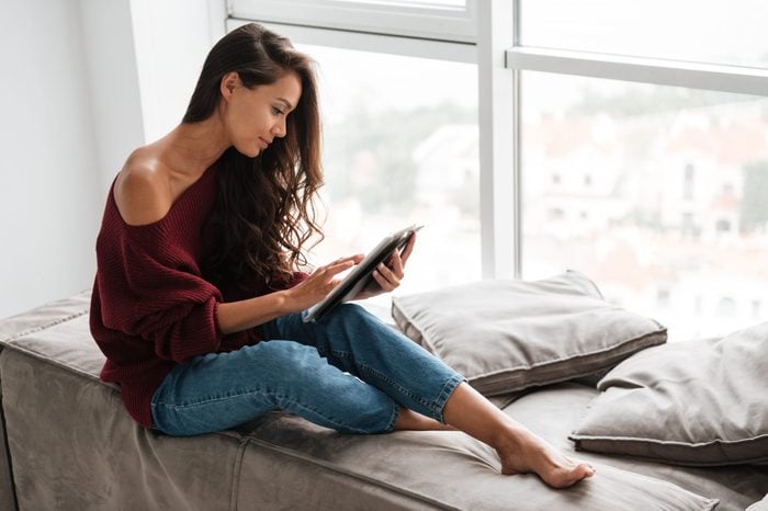 Smiling pretty woman in sweater using tablet computer while sitting on a windowsill at home