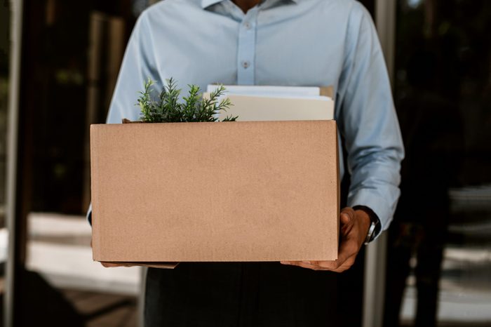 Close up of box with working stuff including pot plant and documents. Fired man is standing outside and holding all his things after leaving company