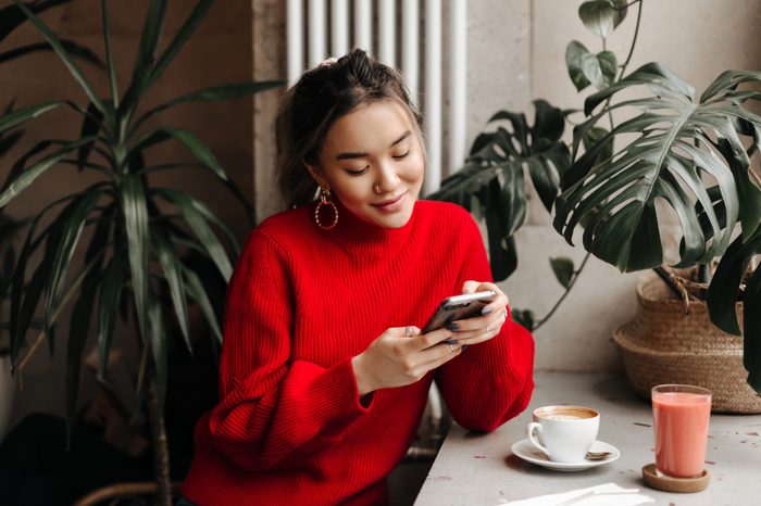 Charming young girl in massive earrings and bright sweater chats in phone while sitting in cafe over cup of coffee