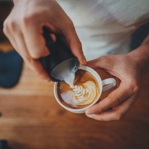 Top view shot of professional barista pouring milk from jar in to a cup of coffee, coffee being prepared by a barista. Focus on hands holding cup of coffee