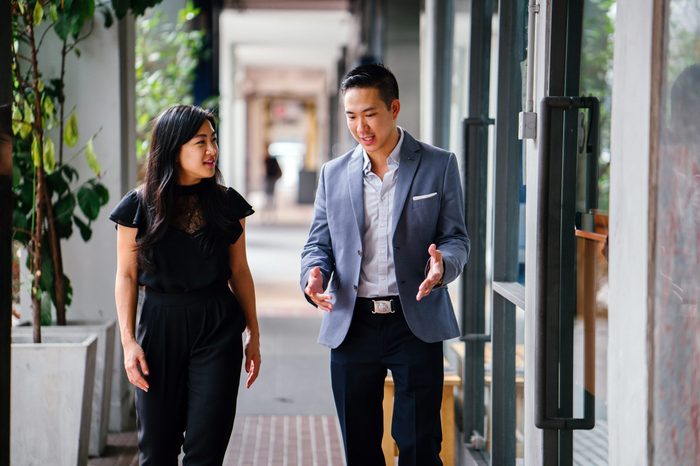 A portrait of two professional business people walking and talking. They are deep in conversation as they walk on a street in a city in Asia. The man and woman are both professionally dressed. 
