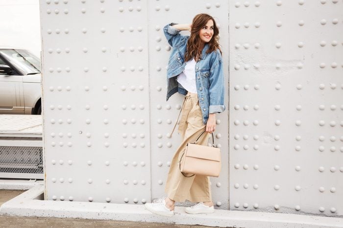 Outdoors fashion portrait of trendy pretty girl posing on the white wall background. Smiling and walking on the city. Going shopping. Wearing stylish wide jean jacket, beige slacks. Positive emotione