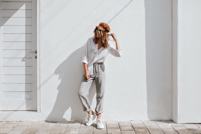 Stylish girl in gray pants and white cotton blouse posing near white wall. Photo of woman in cap and glasses