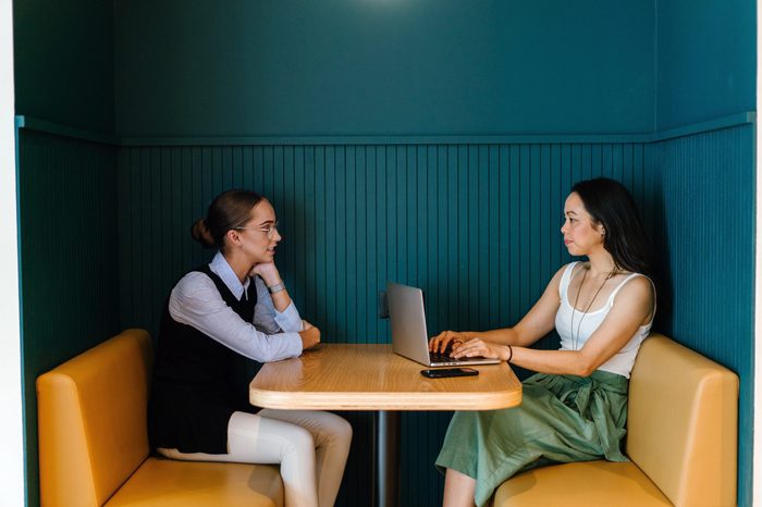 A Caucasian and Asian woman sit in a trendy booth in an office and have a business discussion in the office. They are both young, attractive and are having a focused talk about work. 