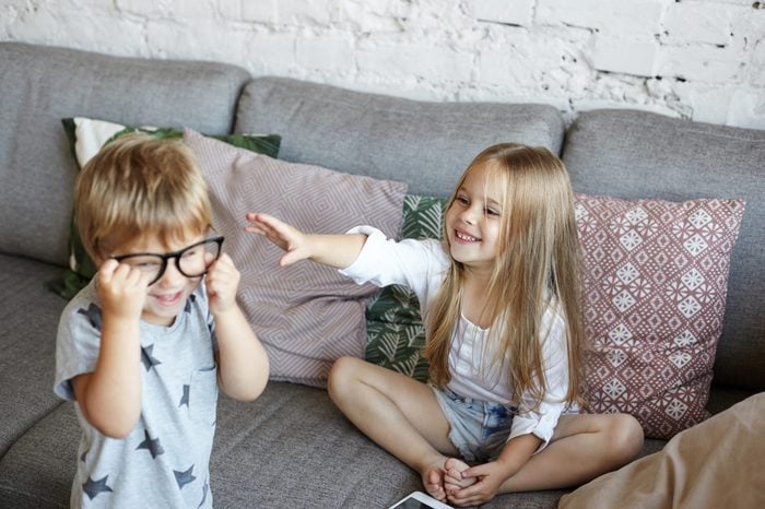 Little sister and brother having fun indoors after kindergarden: cute girl sitting on sofa and reaching out hands to her brother who wearing his father's glasses. Childhood, leisure and fun concept