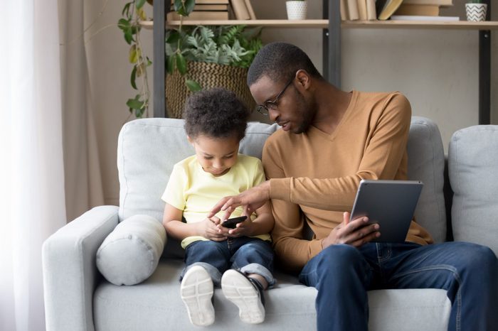 African father and son sitting on sofa in living room use tablet and smartphone spends time at home with electronic devices, dad helps explains to kid new app or game. Bad habit, wireless tech concept