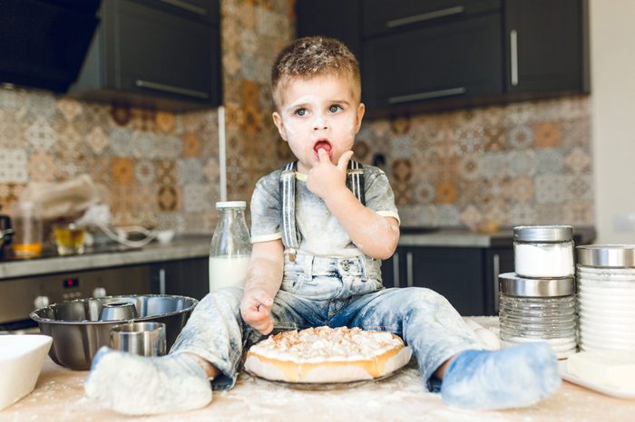 Funny kid sitting on the kitchen table in a roustic kitchen playing with flour and tasting a cake. He is covered in flour and looks funny. He puts his finger in the mouth.