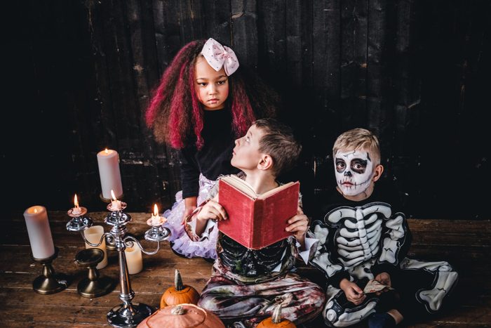 Group of cute multiracial kids in scary costumes reading horror stories in an old house, during Halloween party. 