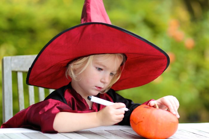 Portrait of little girl in Halloween witch costume decorating pumpkin outdoors in the garden. Kraft for kids.