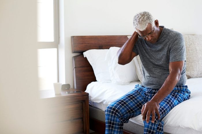Senior Man Suffering With Neck Pain Sitting On Side Of Bed At Home