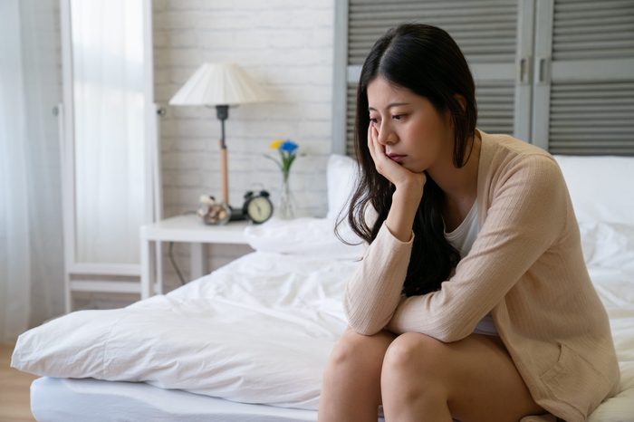 Depressed young girl in casual wear sit on bed in cozy bedroom. thoughtful asian woman resting at home put hand on chin thinking problems. worried lady feeling sad indoors on weekend looks frustrated