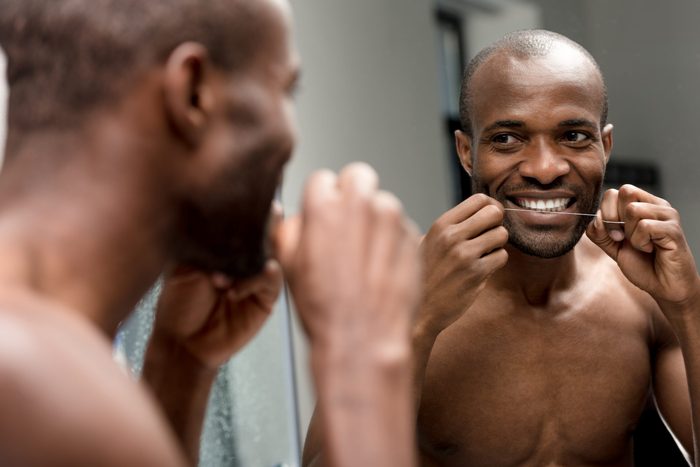 smiling young african american man holding dental floss and looking at mirror in bathroom
