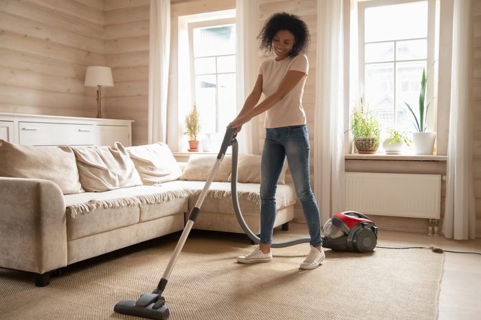 Full length positive 30s mixed race woman enjoy weekend time at modern cozy apartments, girl using vacuum cleaning carpet in living room at sunny warm day, Housekeeping chores and home routine concept