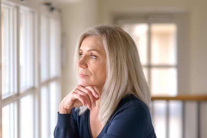 Attractive thoughtful woman with serious expression standing with her hand to her chin staring quietly out of a large window