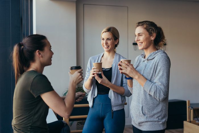 Women relaxing after workout holding coffee and talking in a pilates gym.