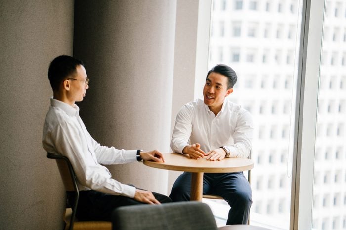 Two Chinese Asian business men have a chat at a table in the office in a meeting room during the day. They are professionally dressed in shirt and pants and having a focused discussion together.
