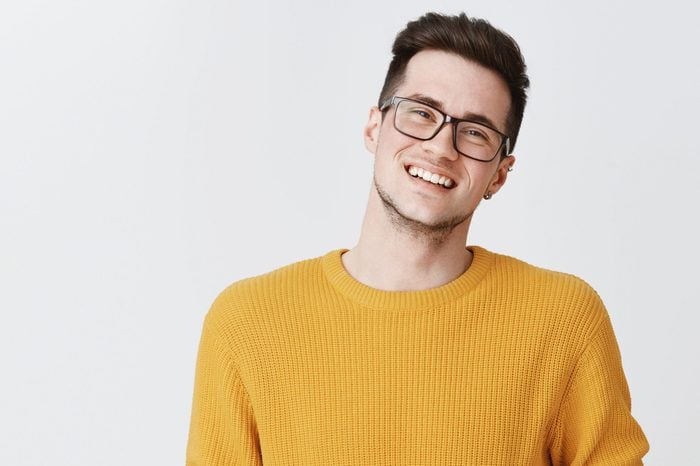Waist-up shot of happy and delighted handsome young man in glasses and yellow sweater tilting head, smiling and laughing as looking friendly at camera on right side of copy space over gray background
