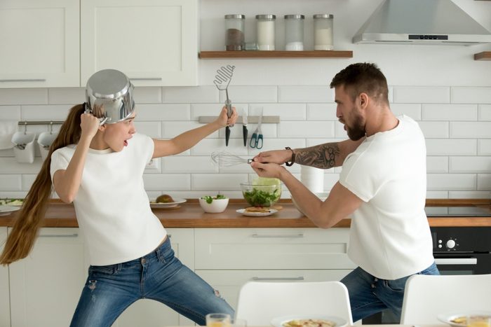 Funny couple pretending fight with utensils tools while cooking at home together, husband and wife having fun feeling playful holding kitchenware struggling in the kitchen preparing healthy food
