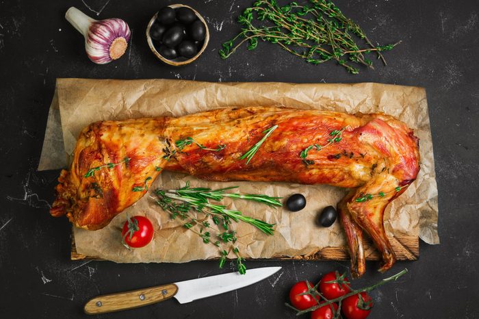 Roast baked whole rabbit on wooden board on paper. Ingredients for baked rabbit rosemary, thyme, cherry tomatoes, garlic, olives on dark background. A festive meal. Top view. 