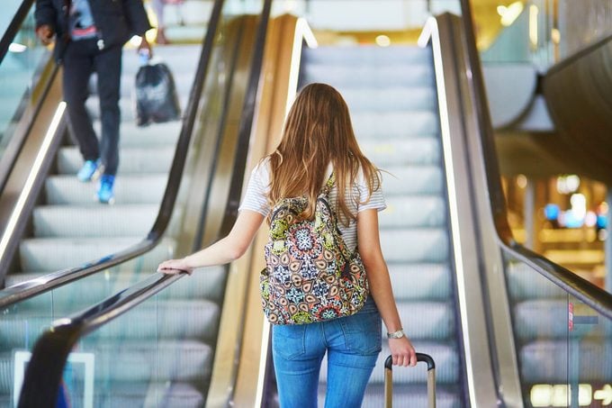 Beautiful young tourist girl with backpack and carry on luggage in international airport, on escalator
