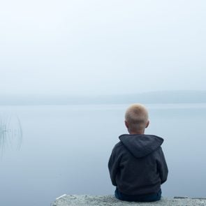 sad child sitting alone by lake in a foggy day, back view