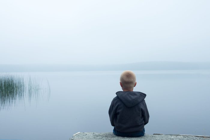 sad child sitting alone by lake in a foggy day, back view