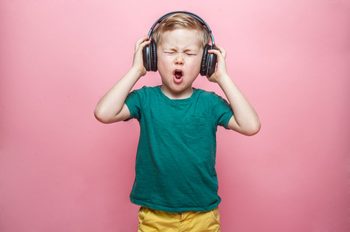 Stylish teen boy listening music in headphones and singing against pink background. School child listening loud music in wireless earphones and dancing.