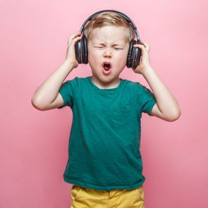 Stylish teen boy listening music in headphones and singing against pink background. School child listening loud music in wireless earphones and dancing. 