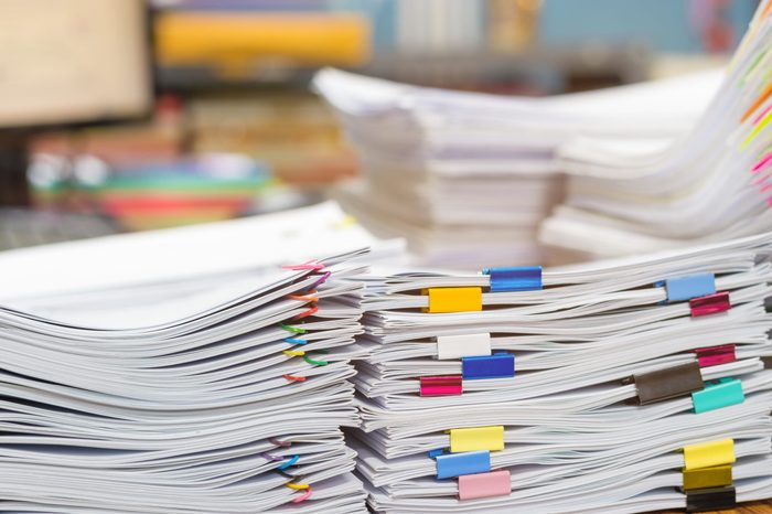 Pile of unfinished document on office desk. Stack of homework assignment archive with colorful paper clip and binder clip on teacher table waiting to be managed. Education and business concept.