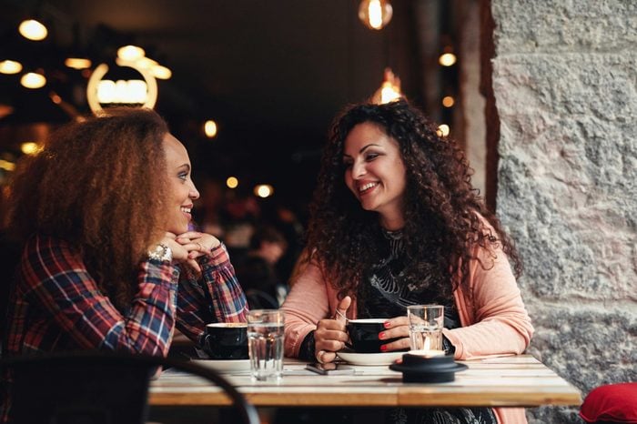 Two women discussing at a restaurant smiling. Young friends sitting in a cafe and talking.