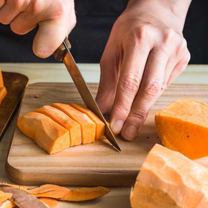 Young woman cutting with knife sweet potato into wedges, peels on wood table, sliced carrots, kitchen interior, close up