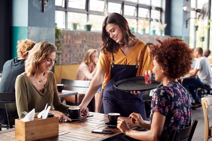 Two Female Friends Sitting At Table In Coffee Shop Being Served By Waitress