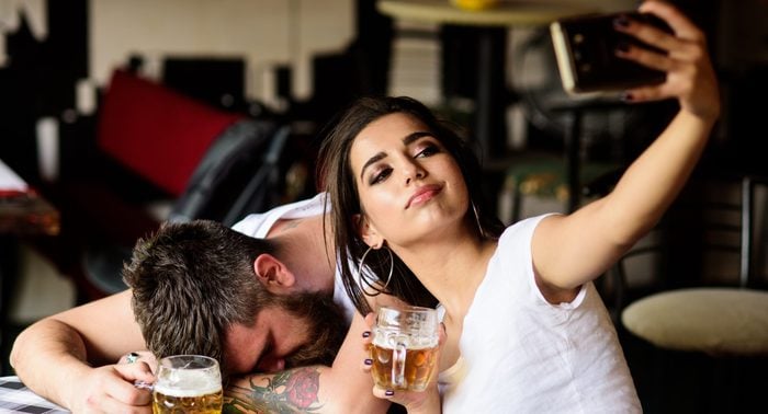 Take selfie to remember great event. Woman making fun of drunk friend. Man drunk fall asleep table and girl with full beer glass. Girl taking selfie photo drunk boyfriend. He appears too weak for her.
