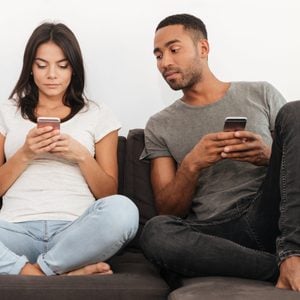 Photo of young couple using their cellphones sitting on sofa at home. Boy looking at woman's phone.