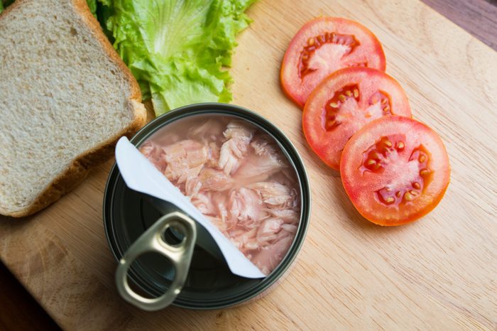 Preparing tuna sandwich, tuna in can opened with bread, lettuce, and tomato on wood background