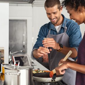 Young man cooking with girlfriend and adding spice to the sauce. Guy adds black pepper into frying pan on stove while woman using spatula to mix. Multiethnic couple preparing lunch together at home.