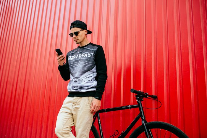 Concentrated trendy young man using a mobile phone, browsing websites, sitting on bike. Outdoors. Wearing stylish clothes, sunglasses, in cap.