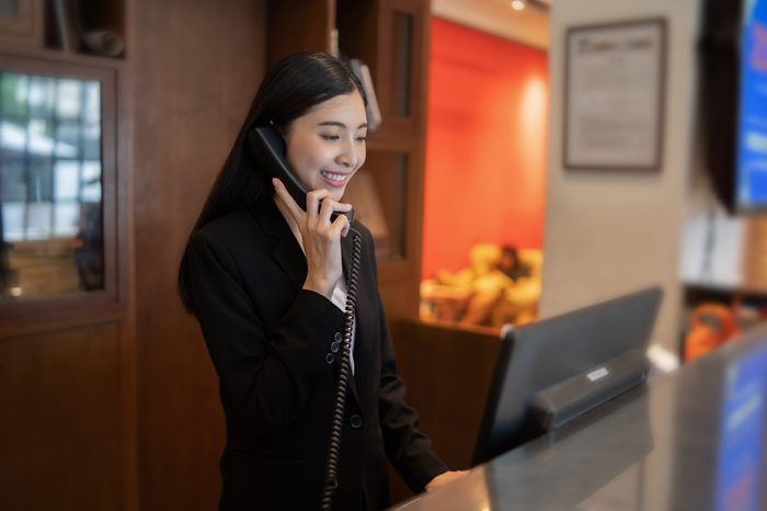 Welcome to the hotel,Happy young Asian woman hotel receptionist worker smiling standing,she taking telephone call at a Modern luxury reception counter waiting for guests getting key card in hotel 