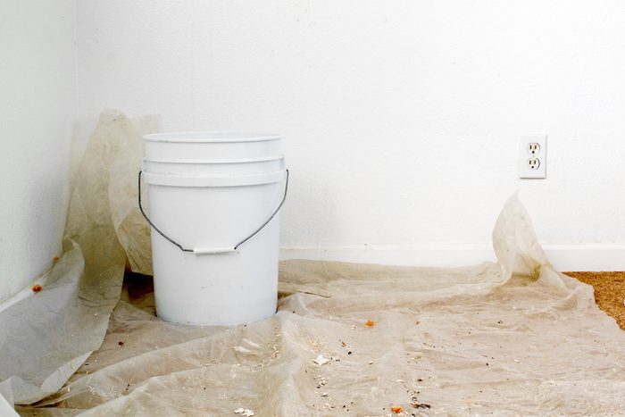 A white bucket over a sheet of plastic placed over carpet floor to collect water from leaking ceiling