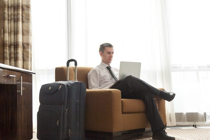 Portrait of handsome young business man wearing formal white shirt and black tie sitting in armchair in hotel room with packed luggage bag on the floor. Traveller working on laptop