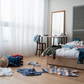 straw hat and colorful clothes in luggage on wooden floor. empty nobody in messy white bed in bedroom packing suitcase for travel abroad summer vacation holidays. mirror by window at dressing table.