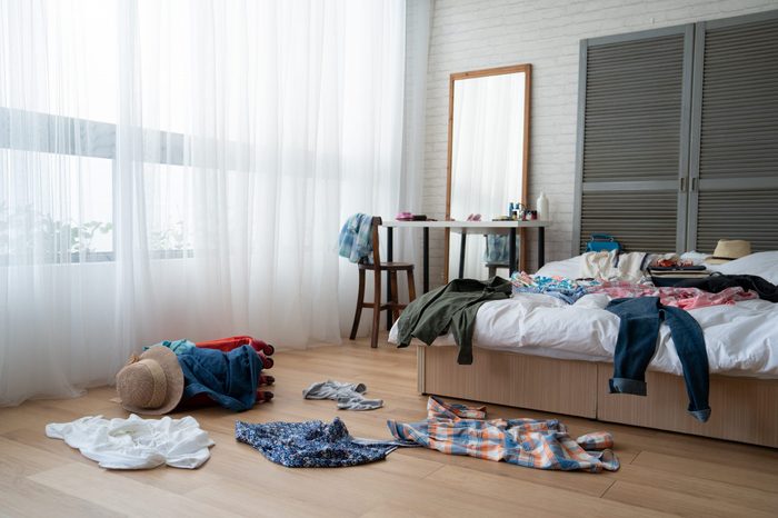 straw hat and colorful clothes in luggage on wooden floor. empty nobody in messy white bed in bedroom packing suitcase for travel abroad summer vacation holidays. mirror by window at dressing table.