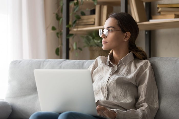 Dreamy millennial woman sit on couch hold laptop look in distance thinking distracted from online work, thoughtful girl in glasses take break from computer studying dreaming or visualizing at home