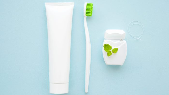 Toothbrush with green bristles, white tube of toothpaste, container of dental floss and peppermint leaf on pastel blue background. Fresh breath and healthy teeth concept.