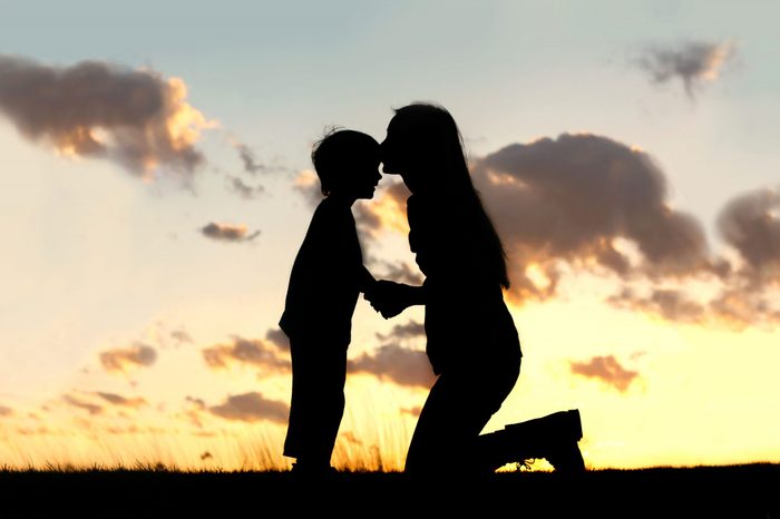 Silhouette of a young mother lovingly kissing her little child on the forehead, outside isolated in front of a sunset in the sky on a summer day.