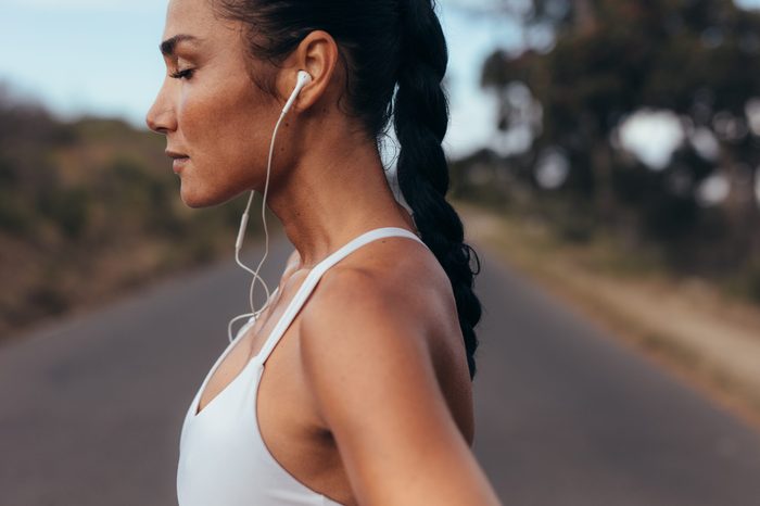 Side view of a female runner standing on a road in morning. Woman in fitness gear standing on a road listening to music using earphones.