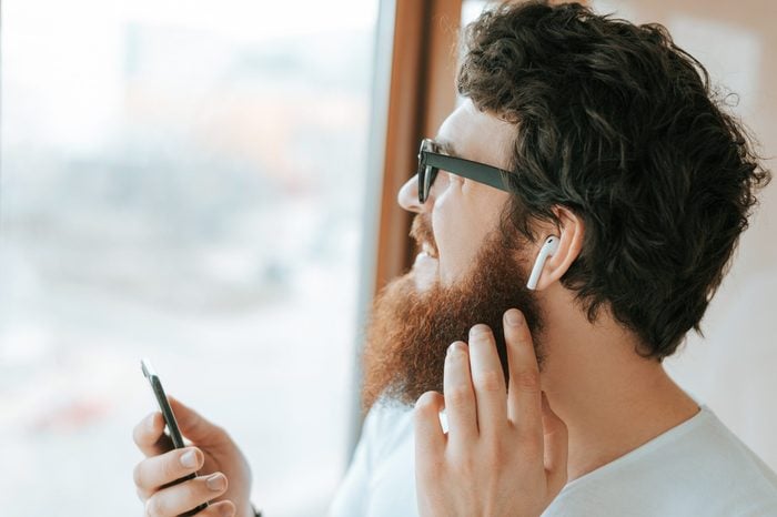 Handsome bearded businessman is using mobile and airpods, while talking with coworker near window