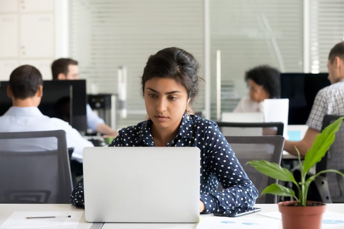 Focused indian female employee busy working online at laptop in shared office, concentrated millennial girl intern using computer in coworking space, prepare project or surfing internet