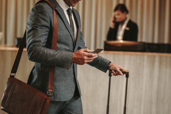 cropped shot of businessman in hotel lobby with mobile phone and luggage. Male business traveler arriving at his hotel, with focus on hands holding smart phone.